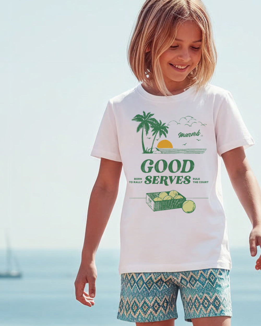 Child wearing a white t-shirt with a tennis graphic on the front and patterned shorts, standing near the seaside.