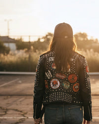 Person wearing a black leather jacket with patches and studs, standing in an abandoned tennis court at sunset.