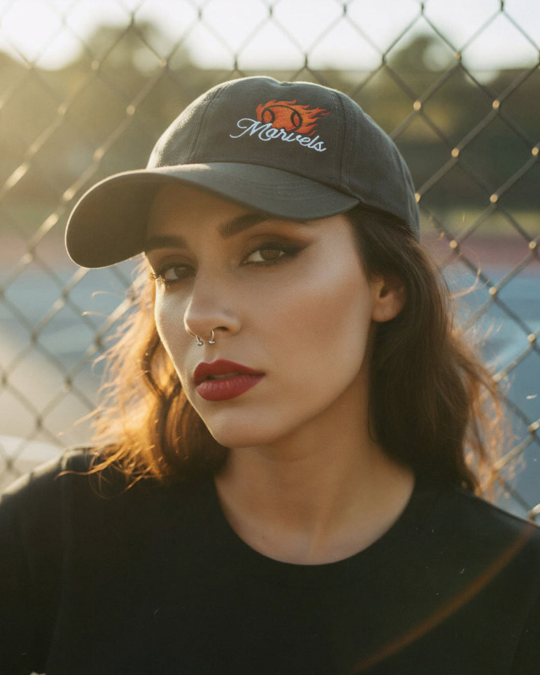 Woman wearing a cap with 'Marvels' logo in front of a chain-link fence.