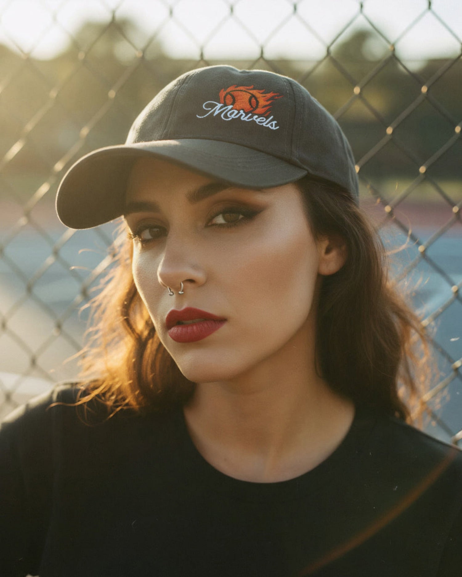 Woman wearing a cap with 'Marvels' logo in front of a chain-link fence.