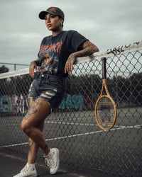Person leaning against a tennis court fence with a racket, wearing a graphic t-shirt and cap.