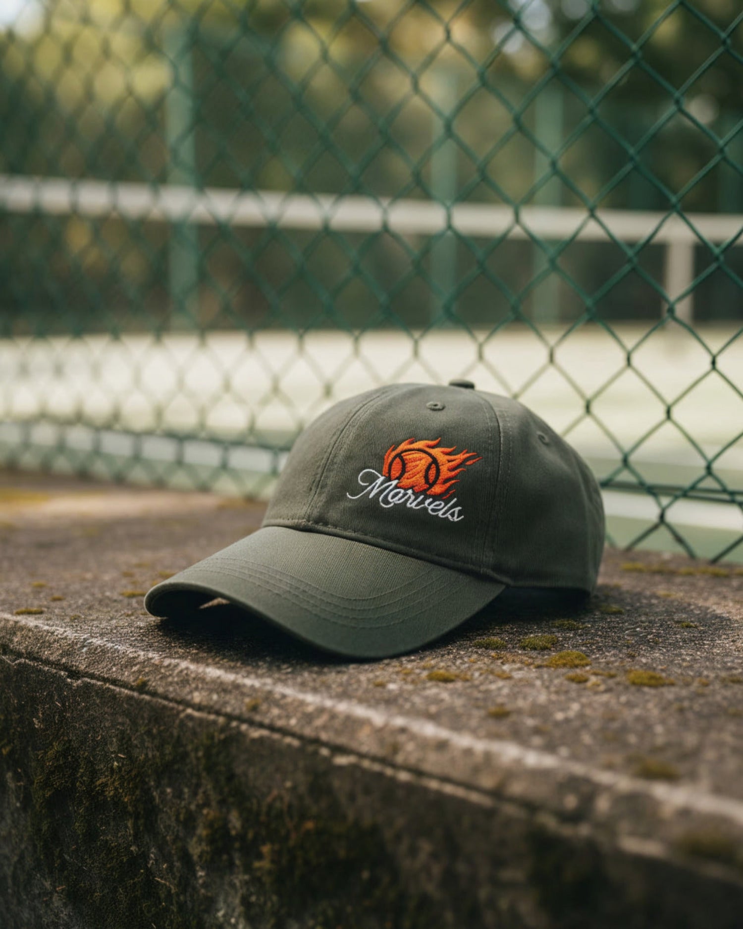 Green cap with a logo on a concrete surface with a chain-link fence and tennis court in the background