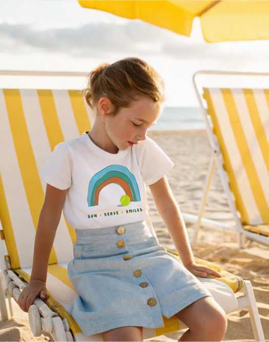 Kid on a beach wearing the Sun Serve Smiles rainbow tennis t-shirt