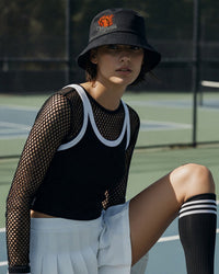 Person wearing a black bucket hat, black mesh top, white shorts, and black knee-high socks with a tennis court background.