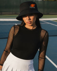 Person wearing a black bucket hat and black top with fishnet sleeves on a tennis court
