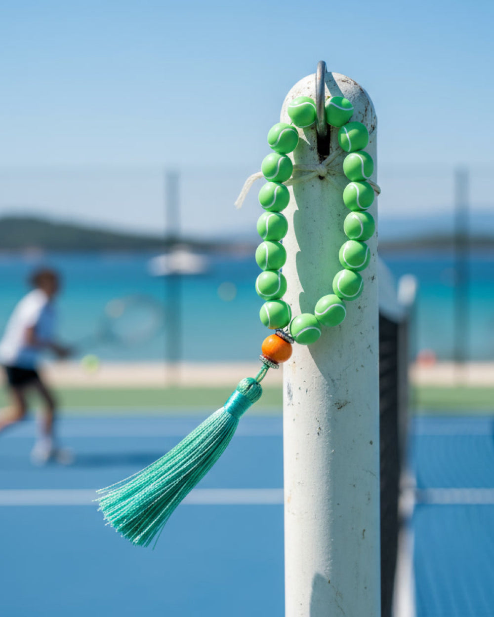 Green tennis ball beaded tespih with a tassel on a tennis court post, with a blurred tennis player in the background.