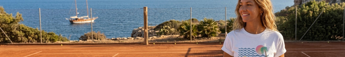 A woman wearing a white t-shirt with a colorful graphic print stands on a clay tennis court with a scenic coastal background featuring blue water, rocks, and a sailboat.
