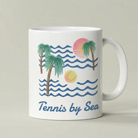 A white ceramic coffee mug with a colorful retro-style graphic of palm trees, blue ocean waves, a pink and orange sun, and a yellow tennis ball, with the text 'Tennis by Sea' printed below.