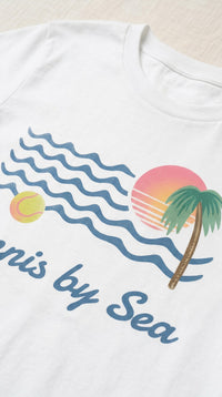 White short-sleeve t-shirt featuring a retro beach graphic with blue waves, a pink and orange sunset, a palm tree, and the text 'Tennis by Sea' in blue script.
