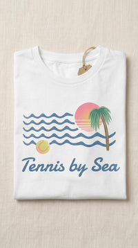 A folded white t-shirt with a retro beach graphic that reads 'Tennis by Sea'. The graphic features blue wavy lines for the ocean, a pink and orange sunset, a green palm tree, and a yellow tennis ball.