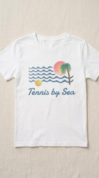 A white short-sleeve crewneck t-shirt laid flat on a light beige textured fabric background. The shirt features a retro graphic of blue ocean waves, a yellow sun, a pink and orange sunset, a green palm tree, and the text 'Tennis by Sea' in blue script.