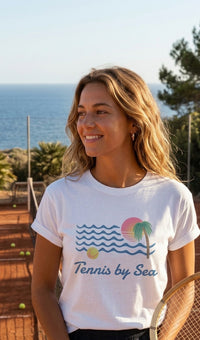 A woman wearing a white graphic t-shirt with a 'Tennis by Sea' design, standing on a tennis court with the ocean in the background.