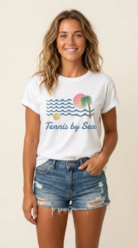 A woman modeling a white graphic t-shirt with a 'Tennis by Sea' design featuring blue waves, a green palm tree, and a pink sun, paired with distressed denim shorts against a light beige background.