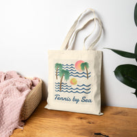 Tote bag with a tennis and waves design on a wooden surface with a white wall and plant in the background.