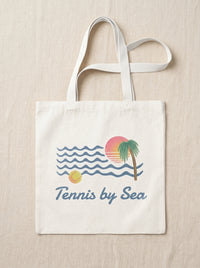 A cream-colored canvas tote bag with white shoulder straps, featuring a retro-style graphic of blue waves, a yellow tennis ball, a green palm tree, and a pink/orange sunset, with the text 'Tennis by Sea' printed below. The bag is displayed flat against a neutral beige linen background.