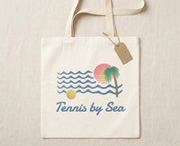 A cream-colored canvas tote bag with a retro beach-themed graphic that reads 'Tennis by Sea'. The design features blue ocean waves, a pink and orange sunset, a green palm tree, and a tennis ball.