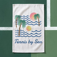A white cotton tea towel featuring a retro beach graphic with palm trees, blue waves, a pink and orange sun, and a yellow tennis ball, with the text 'Tennis by Sea' printed below. The towel is displayed flat against a green tennis court background with white lines.