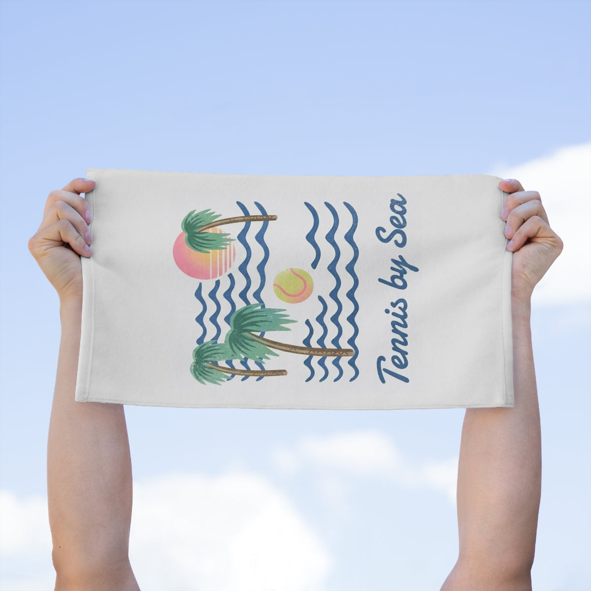 A person holds up a white-colored towel featuring a tropical beach illustration with palm trees, blue waves, a pink and yellow sun, and the text 'Tennis by Sea' against a light blue sky background.