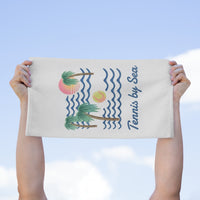 A person holds up a white-colored towel featuring a tropical beach illustration with palm trees, blue waves, a pink and yellow sun, and the text 'Tennis by Sea' against a light blue sky background.