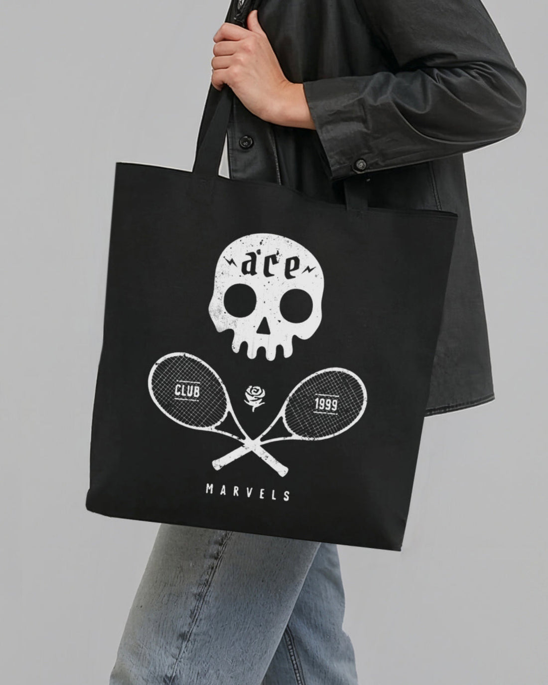 Person holding a black tote bag with a white skull and tennis racket design on a gray background