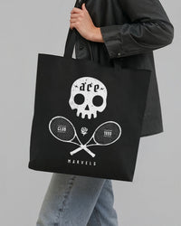 Person holding a black tote bag with a white skull and tennis racket design on a gray background