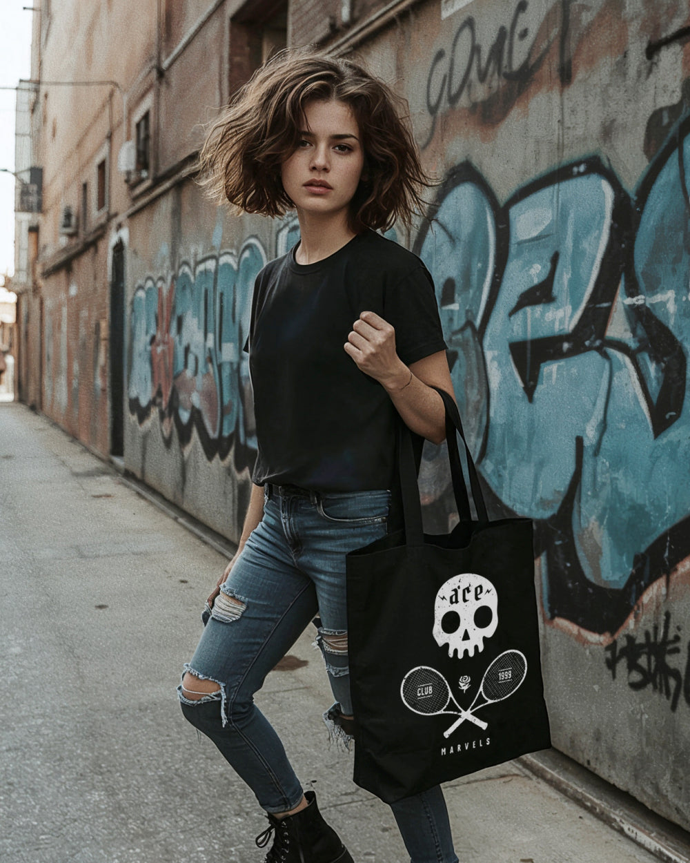 Woman holding a black tote bag with a skull and tennis design against a graffiti-covered wall.