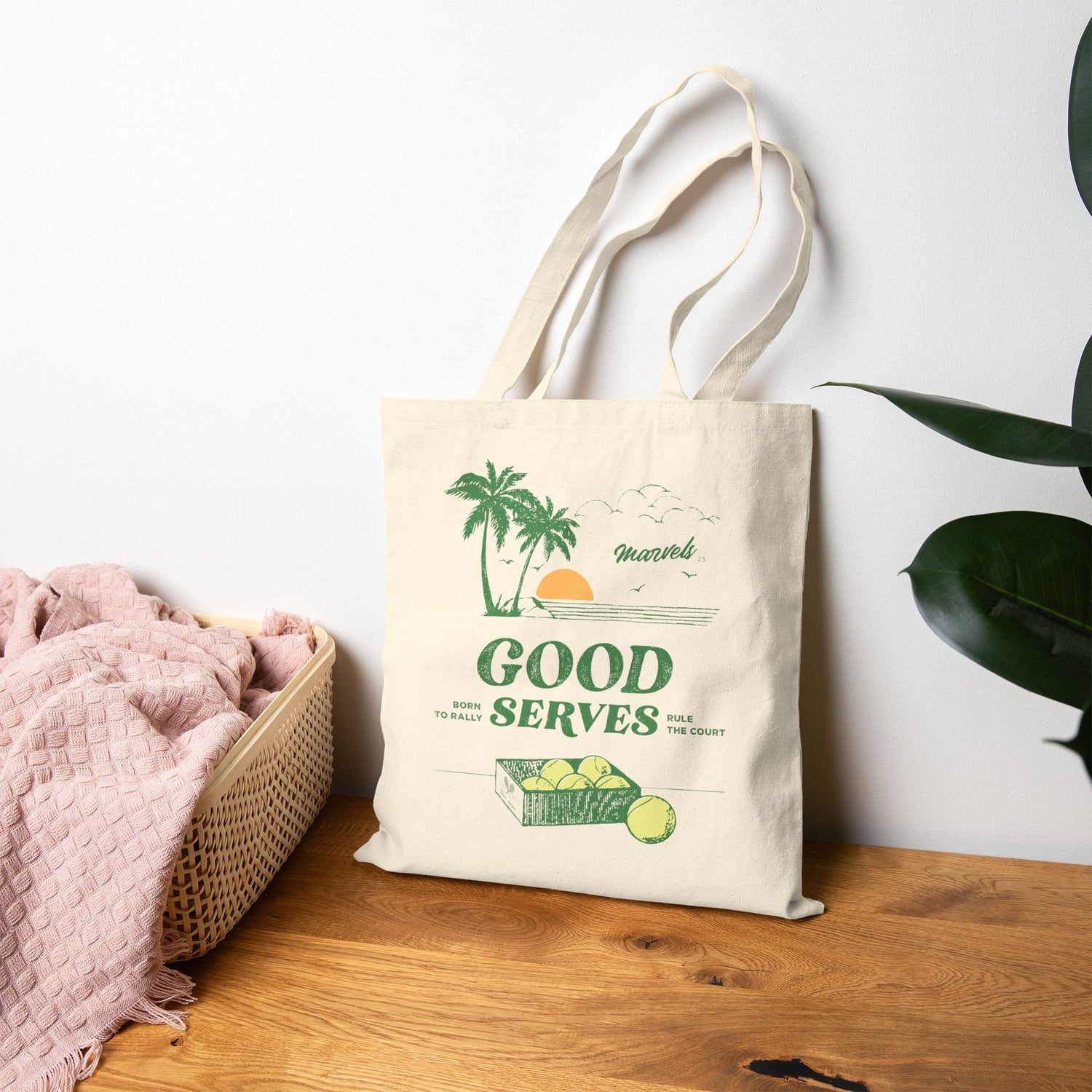 Natural tote bag with green 'Good Serves' text and palm tree graphics on a wooden surface