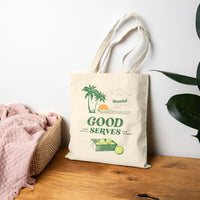 Natural tote bag with green 'Good Serves' text and palm tree graphics on a wooden surface