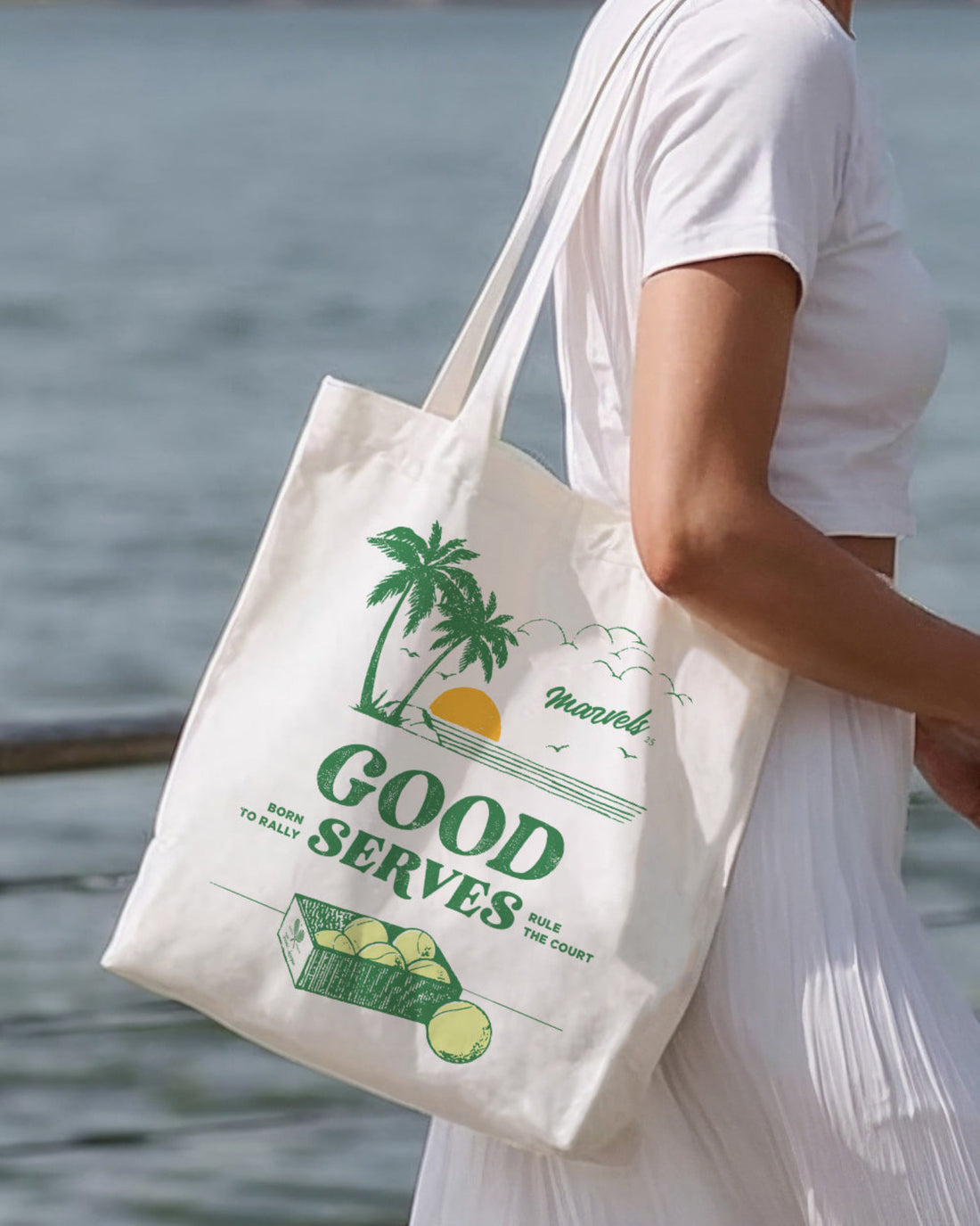 Person holding a tote bag with a palm tree design and 'Good  Serves' text by a body of water.
