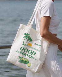 Person holding a tote bag with a palm tree design and 'Good  Serves' text by a body of water.