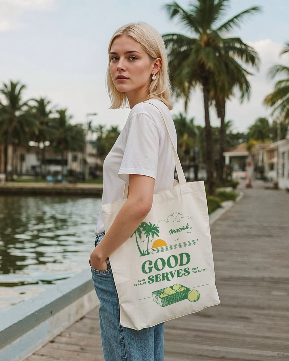 Woman holding a tote bag with a branded Good Serves design by a waterfront with palm trees.