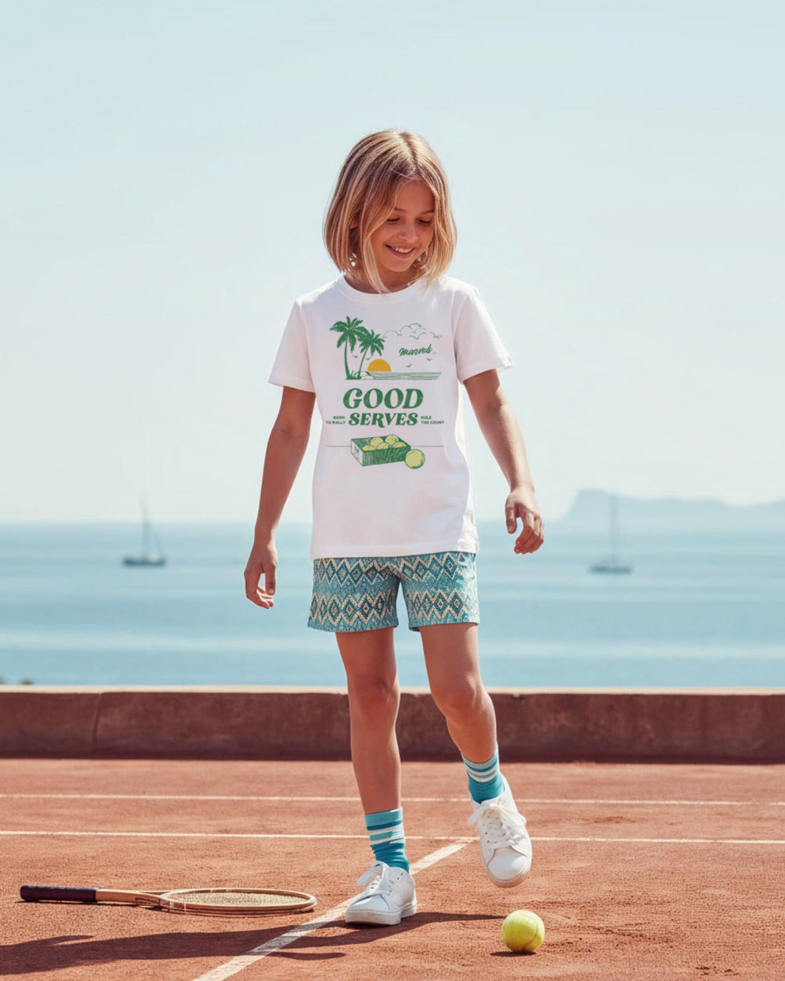 Child standing on a tennis court with a seaside background