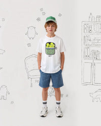 Child wearing a white tennis t-shirt with a graphic design, blue shorts, and a green cap in a room with doodles on the wall.