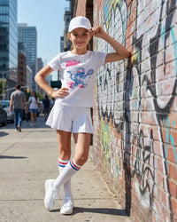 Kid wearing a white t-shirt with a tennis graphic design, white shorts, and white sneakers against a graffiti-covered wall.