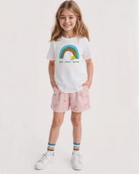 Young girl wearing a white t-shirt with a rainbow design and pink shorts on a white background