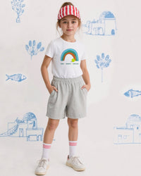 Child wearing a white tennis t-shirt with a rainbow design, gray shorts, and a red and white striped visor