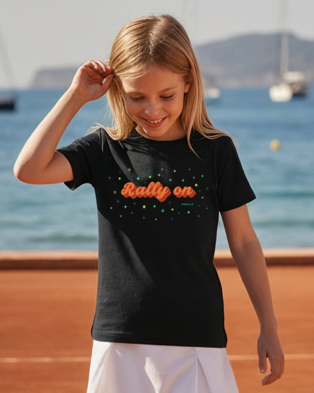Young girl wearing a black t-shirt with 'Rally on' text on a tennis court by the sea.