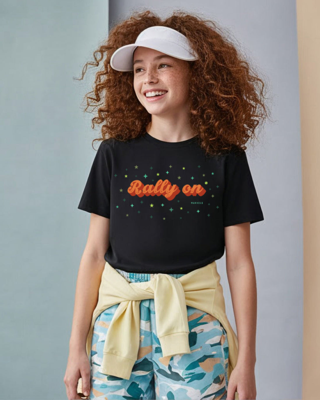 Girl wearing a black t-shirt with 'Rally on' text, colorful pants, and a white cap against a gray background
