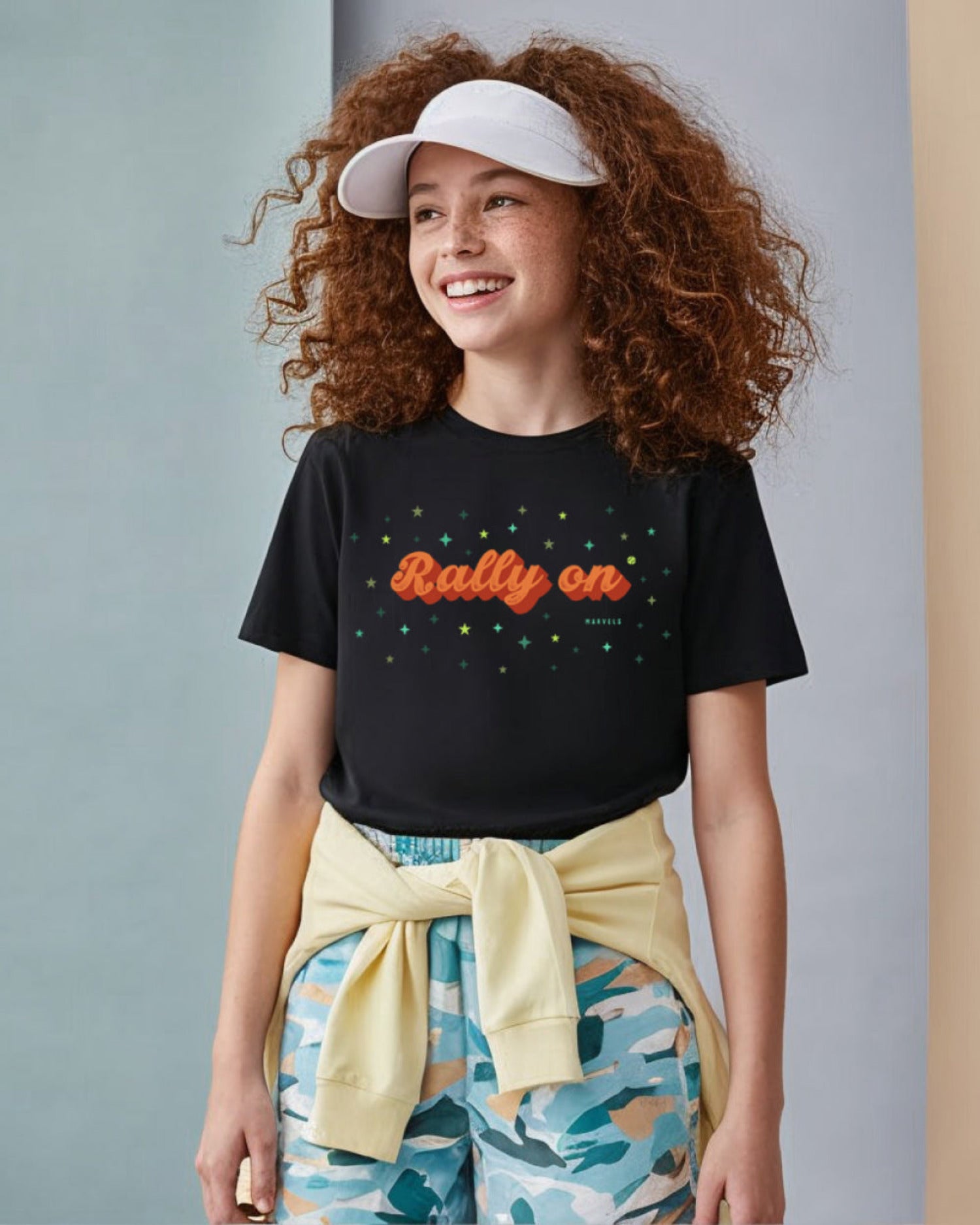 Girl wearing a black t-shirt with 'Rally on' text, colorful pants, and a white cap against a gray background