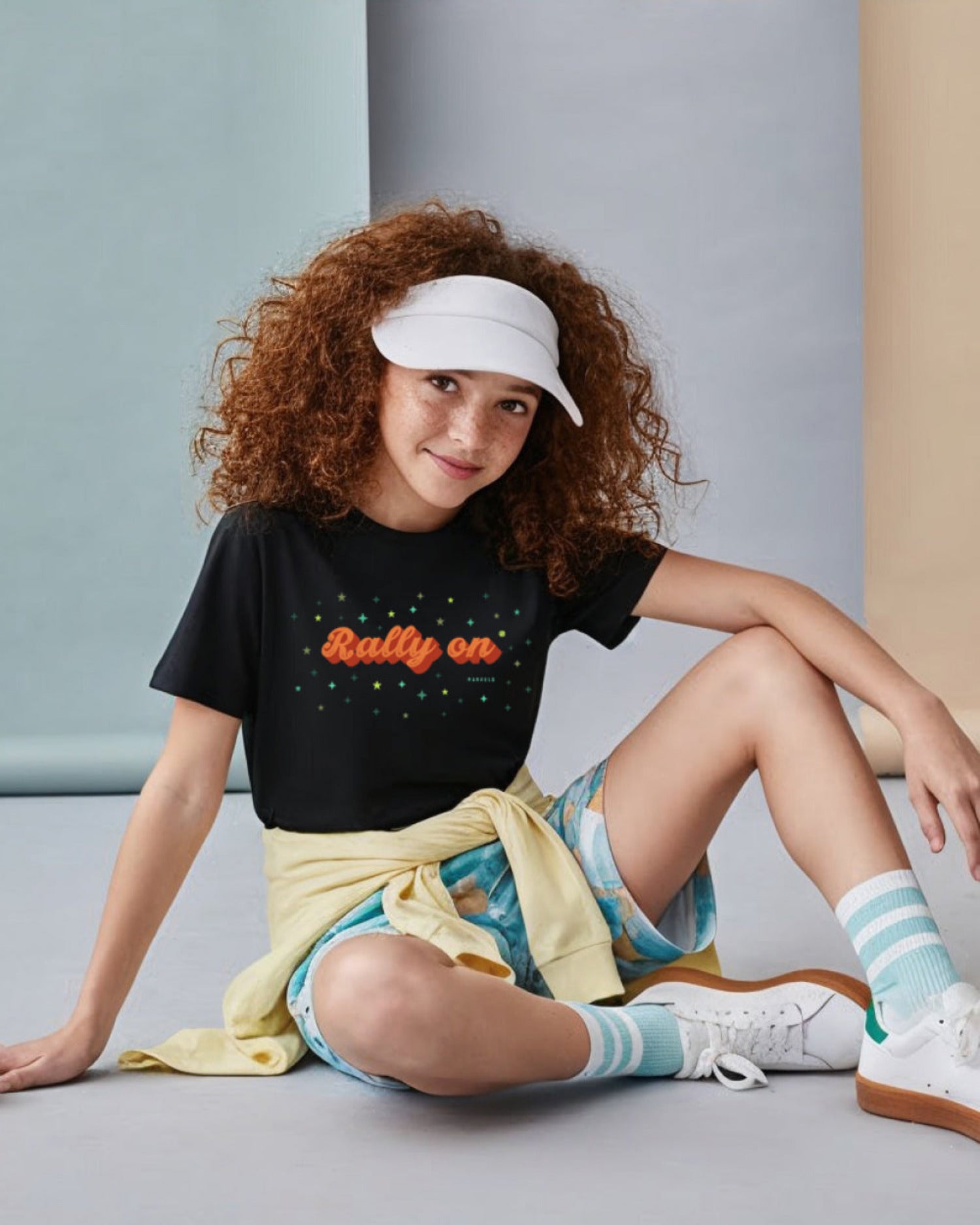 Young girl sitting on a couch wearing a black tennis t-shirt with 'Rally on' text, colorful shorts, and striped socks.