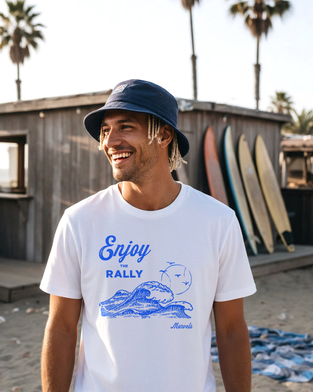 Man wearing a white t-shirt with blue tennis graphics and text, standing in front of surfboards on a beach.