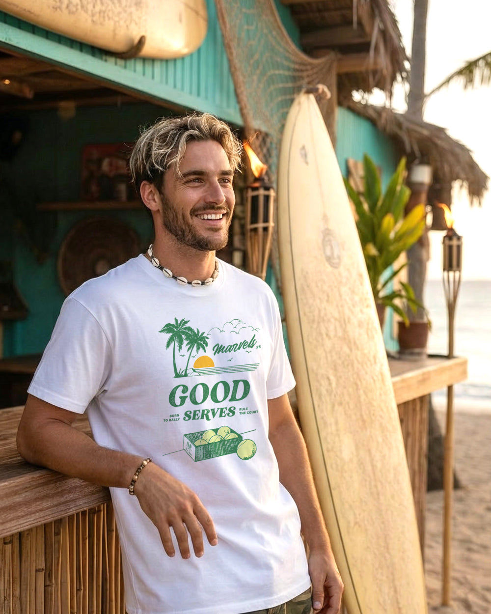 Man wearing a white t-shirt with a tennis graphic design, standing next to a surfboard on a beachside bar.