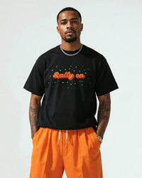 Man wearing a black t-shirt with 'Rally on' text and orange pants against a plain background