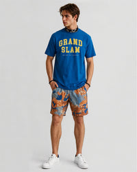 Man wearing a blue 'Grand Slam' t-shirt and patterned shorts on a light gray background