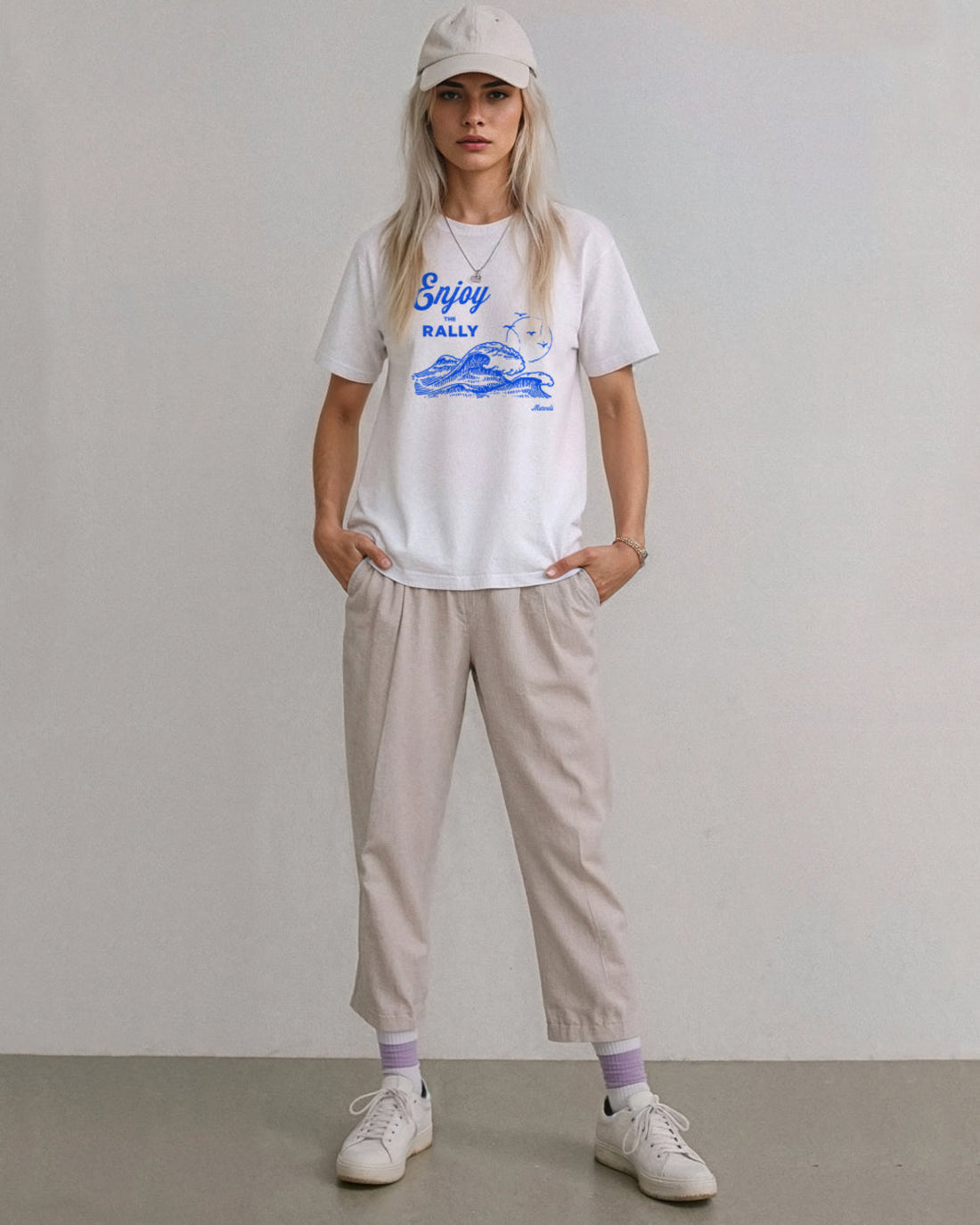 Model wearing a cap, beige pants and a white t-shirt with a blue graphic that reads 'Enjoy the Rally' in text on a plain background