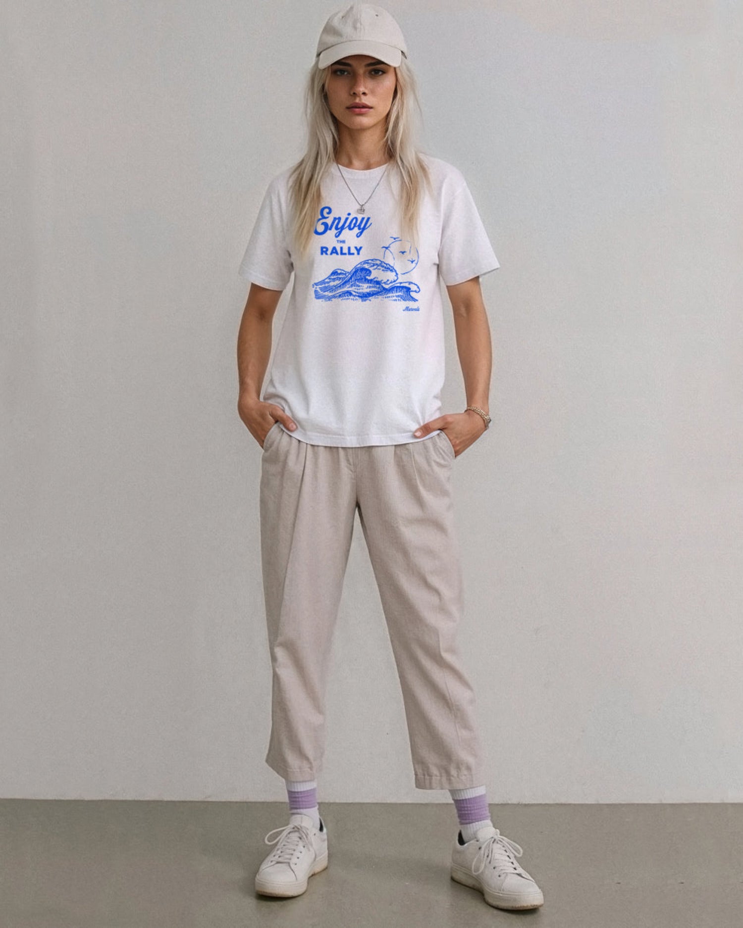 Model wearing a cap, beige pants and a white t-shirt with a blue graphic that reads 'Enjoy the Rally' in text on a plain background