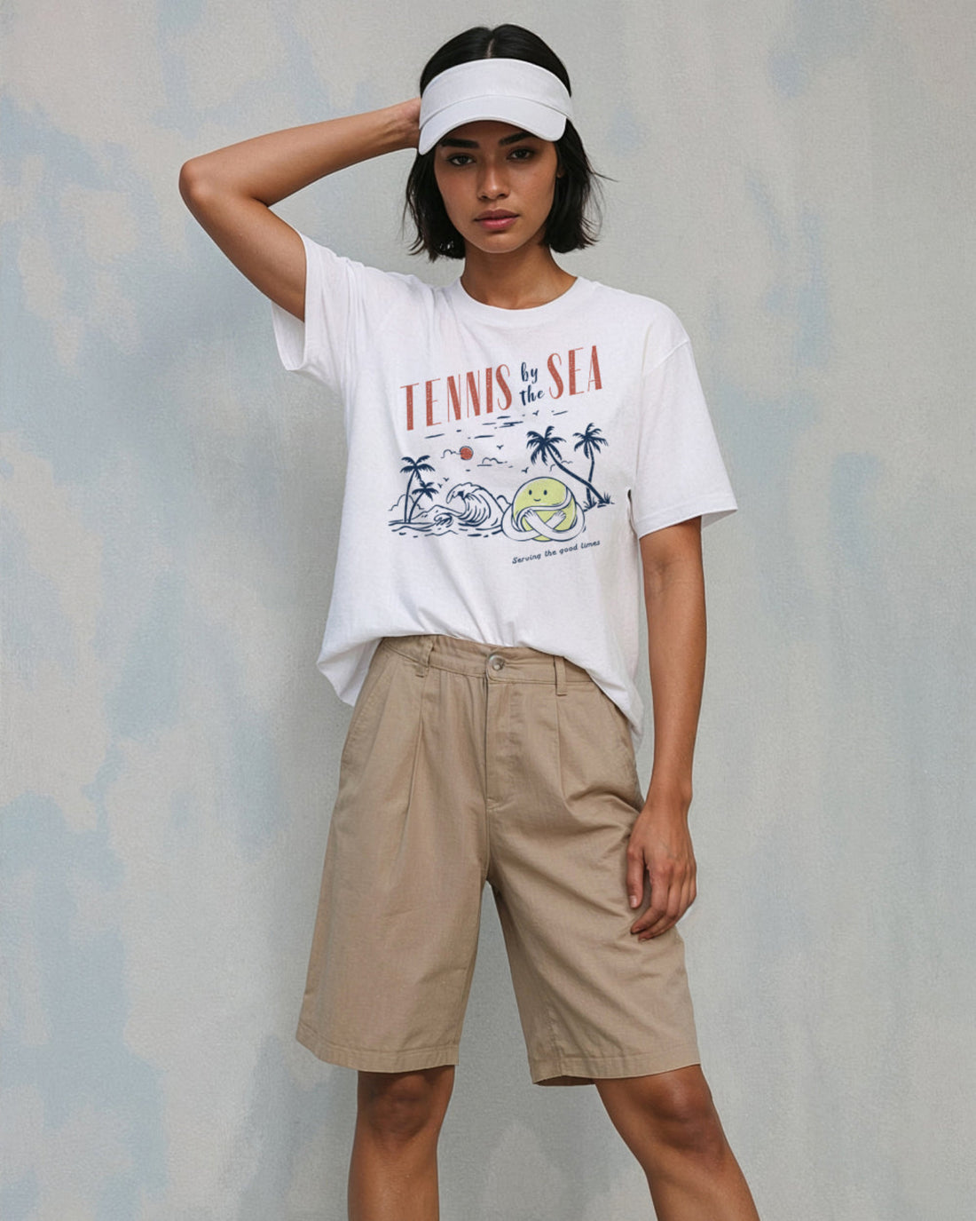 Model wearing beige shorts, a white visor and a white t-shirt with a graphic that reads 'Tennis by the Sea'.