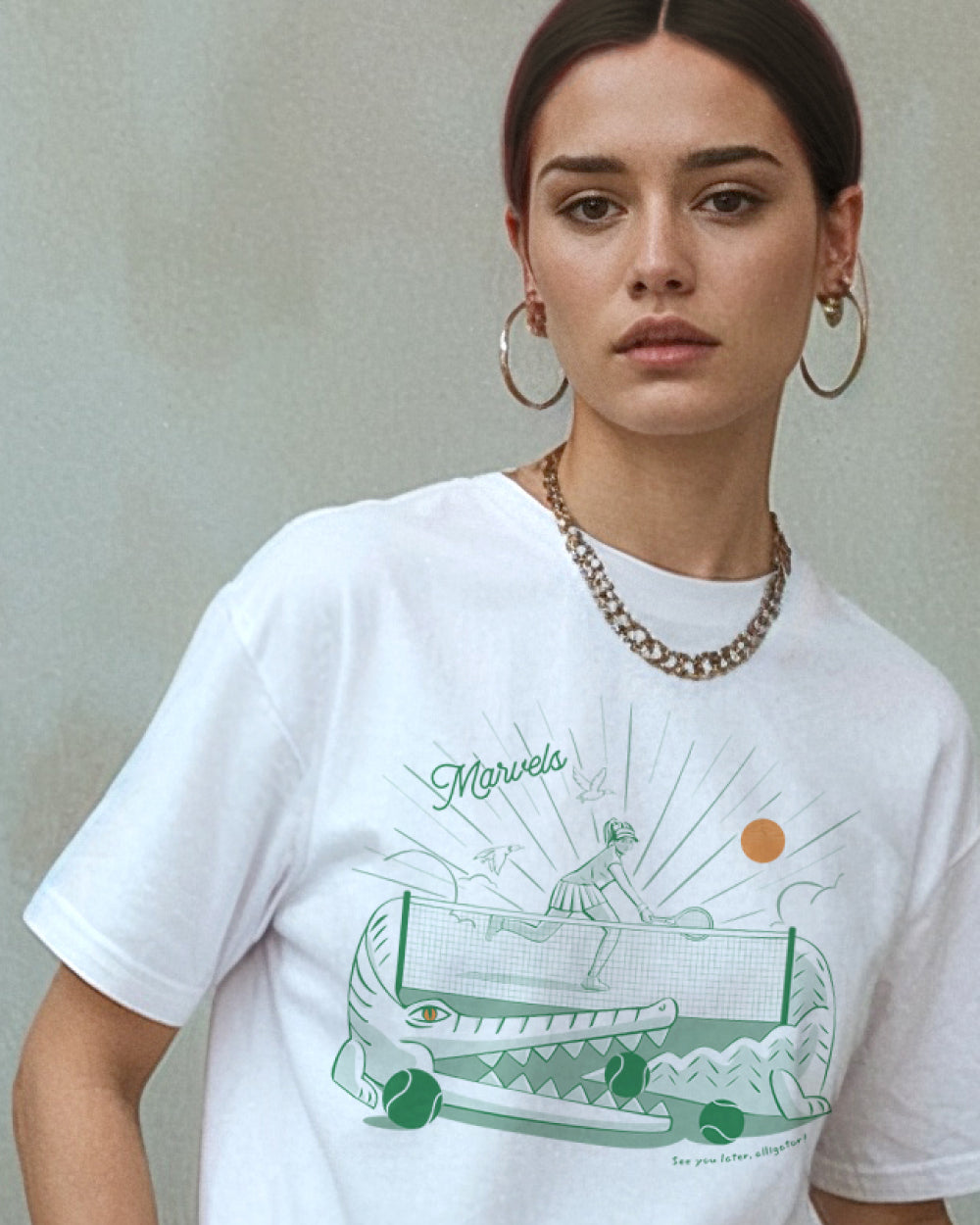 Model wearing a white t-shirt with a tennis graphic with alligator that reads 'See you later' in text on a plain background