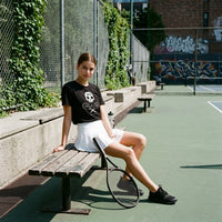 Woman sitting on a bench with a tennis racket on a court, wearing a black t-shirt and white skirt.