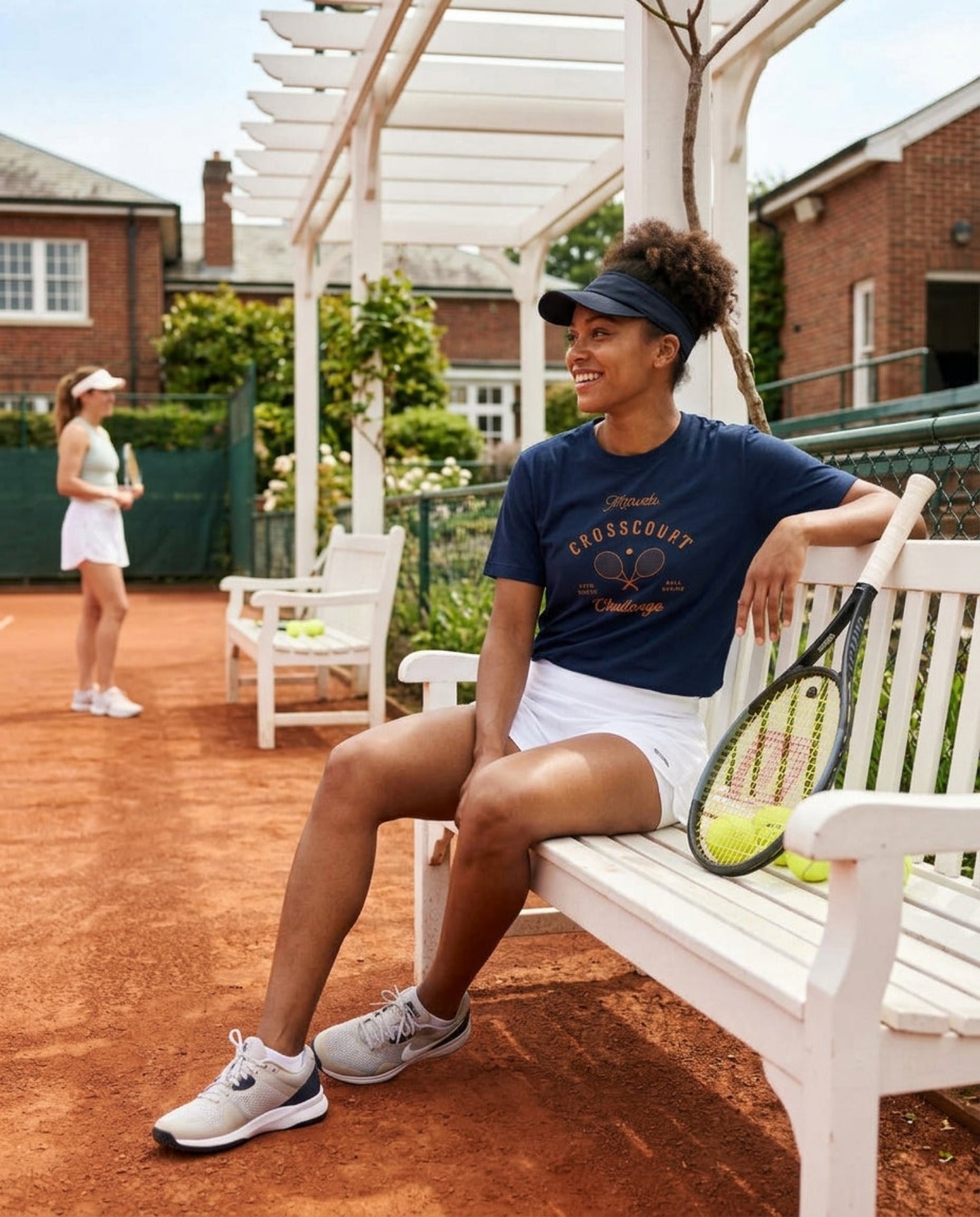 Woman sitting on a bench with a tennis racket on a tennis court, another player in the background.